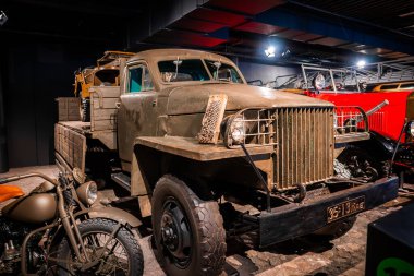 Zeytinli, sıkıcı bir askeri kargo kamyonu kapalı bir yerde klasik bir motosiklet ve kırmızı bir itfaiye arabasının yanında duruyor. Cilalı beton üzerinde spot ışıkları altında, savaş zamanı araç ve zanaatını çağrıştırıyor.