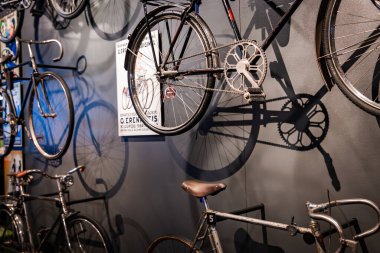 Bir nakliye müzesinde gri bir duvarda çeşitli yüksekliklere çeşitli antika bisikletler monte edilir. Tekerlekler ve zincirler sıcak ışıklar altında gevrek gölgeler bırakır..