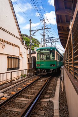 Yeşil Enoden tren vagonu 1201 Kamakura, Japonya 'daki evler arasında hareket ediyor. Üstten elektrik hatları ve geleneksel binalar pisti parlak gündüz ışığında çerçeveliyorlar..