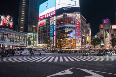 Tokyo 'daki Shibuya Geçidi' nde yayalar bekliyor. Taksi ve arabalar kuyrukta bekliyor. Yüksek binalarda neon panolar parlıyor..