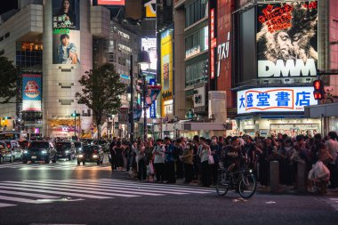 İnsanlar Shibuya 'da, Tokyo yaya geçidinde taksi ve araba kuyruğunda bekliyorlar. Neon reklam panoları arasında DMM panosu ve akşam ışıkları ve yoğun enerjili büyük bir kitapçı tabelası yer alıyor..