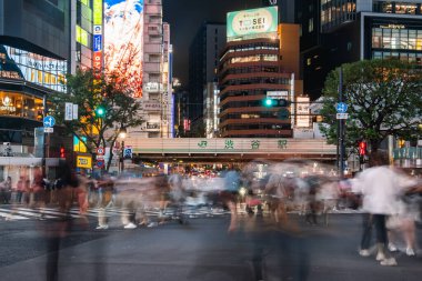 Kalabalık Shibuya 'dan Tokyo' daki JR Shibuya İstasyonu 'nun yakınına akın ediyor. Neon reklam panoları ve uzun pozlama bulanıklığı geceleri kentsel hareket ve enerji gösteriyor.