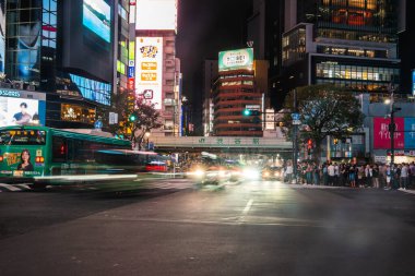 Tokyo 'daki Shibuya Kavşağı' nda insanlar bekliyor. JR köprüsünün altında trafik bulanıklaşırken yeşil ışık, neon panolar parlıyor ve yeşil bir Toei otobüsü kalkıştan önce geçiyor..