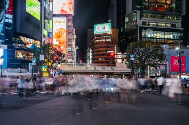 Gece Tokyo 'da, JR Shibuya İstasyonu' nun altında yayalar bulanıklaşıyor, neon panolar parlıyor, uzun pozlar yoğun bir şehir sahnesinde sürekli akış gösteriyor..