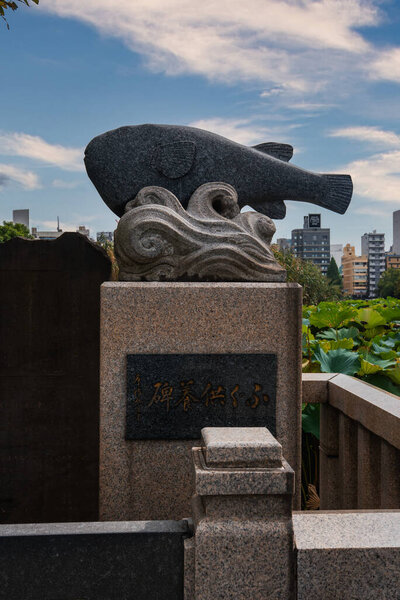 A carved stone fish on waves tops a granite pedestal by a lotus pond in Tokyo, near Shinobazu Pond in Ueno Park, with mid rise buildings visible in soft light.
