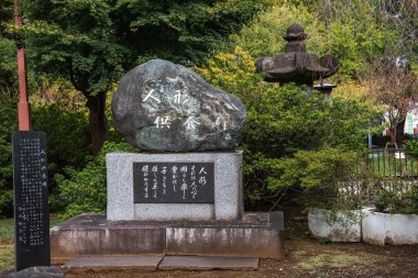 Üzerinde Japon yazıtları olan taş bir anıt, Tokyo 'daki bir parkta, ağaçlar ve çalılarla çerçevelenmiş taş fener ve taşların yanında granitin üzerinde duruyor..