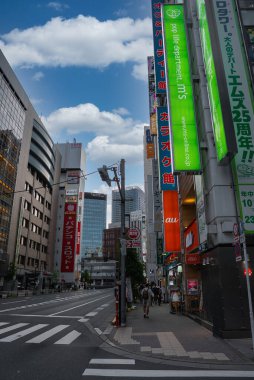 Japon hattında dikey neon ve reklam panolarına sahip uzun binalar bir Tokyo caddesi. Yayalar gün ışığında yürürken Büyük Elma pachinko ve au mobil dükkanı ortaya çıkıyor.