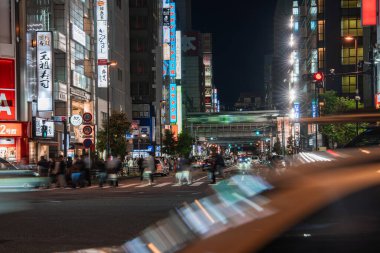 Kalabalık, kanji işaretleri ve neon reklamların altında geceleyin geniş bir Tokyo kavşağından geçiyor. Uzun pozlar araba ışıklarını çizer, yüksek binalar yükselir ve uzak bir üstgeçit yoğun bir sahne oluşturur..