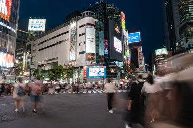 Gece Tokyo 'da Shibuya Geçidi' nden geçen kalabalık, parlak LED reklam panolarının ve kıvrımlı Shibuya 109 cephesinin altında bulanık bir hareketle şehrin nabzını yakalıyor..