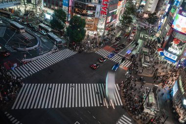 Tokyo 'da Shibuya Scramble Geçidi' nin hava akşam görüntüsü, kalabalık taksi ve otobüsler yoğun kavşaktan geçerken neon panolar parlıyor ve kesişiyor..