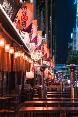 Shibuya ya da Shinjuku yakınlarındaki dar bir Tokyo geçidinde izakaya barları, kanji işaretleri, kırmızı ve beyaz fenerler, noren, boş masalar ve gece yarısı ısıtıcıları görülüyor..