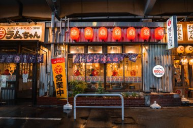 Üstgeçidin altında bir Tokyo izakayası alacakaranlıkta parlıyor, kırmızı fenerler yanıyor, noren asılıyor, yakiniku işaretleri ve görünür içecekler, yağmurlu sokaklar yoğun tabelayı yansıtıyor..