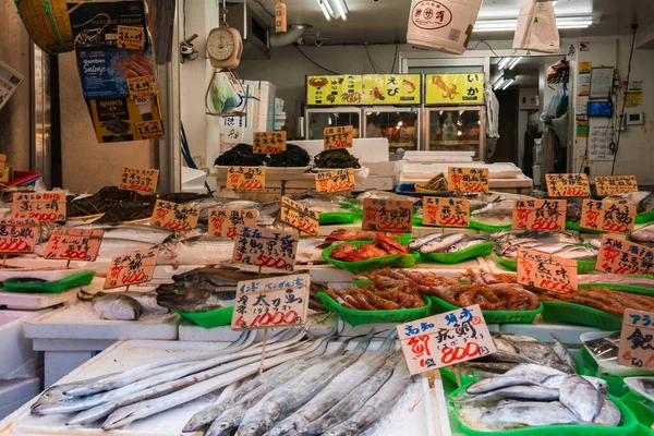 Tokyo pazarında, arka planda Japon fiyat etiketleri, pulları ve asılı tabelaları bulunan yeşil tepsilerde balık, karides, mürekkep balığı ve kabuklu deniz ürünleri sergilenmektedir..