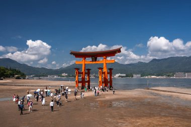 Japonya 'nın Hiroşima kentindeki Itsukushima Tapınağı' ndaki Büyük Torii 'ye sular çekildiğinde kalabalık ıslak kumu geçer. İnsanlar şehrin ve tepelerin ötesindeki tahta sütunları fotoğraflıyor..
