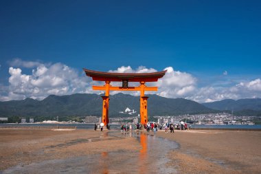 Turistler, Japonya 'nın Miyajima kentindeki Büyük Torii' nin gelgit düzlüklerinde dururlar. Hiroşima Körfezi şehri ve dağları görünür, turuncu kapılar mavi gökyüzüne karşıdır..