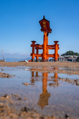 Itsukushima Tapınağı, Miyajima, Hiroşima, Japonya 'daki gelgit düzlüklerinde bir vermilion torii bulunur. Islak kum, taşlar, su birikintisi yansımaları, dağlar ve küçük bir liman görülebilir..
