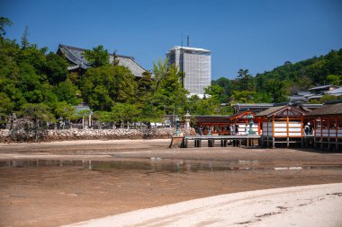 Ziyaretçiler Hiroşima yakınlarındaki Itsukushima Tapınağı 'ndaki gelgit düzlüklerinde orman yamaçları ve görünürde iskele kulesi olan vermilion koridorlar boyunca yürüyorlar.