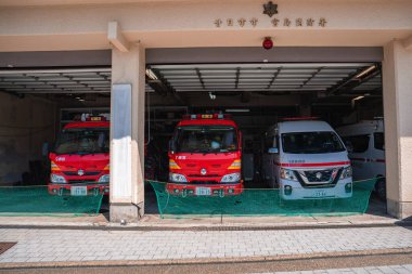 Hiroşima yakınlarındaki Itsukushima Adası 'nda açık bir itfaiye istasyonunda iki kırmızı itfaiye arabası ve beyaz bir ambulans parkı..