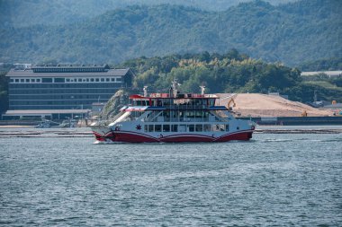 Akçaağaç yaprağı motifli kırmızı ve beyaz bir feribot Hiroşima, Japonya yakınlarındaki sakin bir kanalı geçerek alçak binalar ve görünen ağaçlık tepelerle Itsukushima 'ya gidiyor..