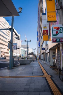 Hiroşima kaldırımında 7 tane 11 işareti, dokunsal sarı şeritler, modern orta katlı binalar ve birkaç yayası ve mağaza önü yakınında bisikleti olan uzak tramvay hatları var..