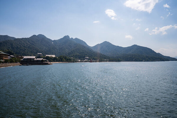 Wide view of the Seto Inland Sea frames Mount Misen on Itsukushima Island near Hiroshima. Gentle waves lead to a coastal settlement with traditional buildings along the shore.
