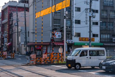 Japonya, Osaka 'da bir demiryolu geçidinde bir kei minibüsü bekliyor. Çizgili bariyerler, tabelalar, dükkan önleri ve kalın kablolar, sabahın geç saatlerinde alçak binalar çerçeveleniyor..