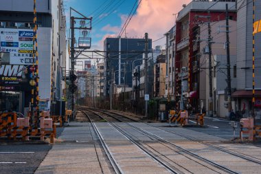 Bir Osaka demiryolu geçidi çizgili bariyerler, işaret lambaları ve kıvrımlı çift raylı teller gösteriyor. Düşük ve orta katlı binalar, sıcak pembe bulutlar ve serin mavi renkler..