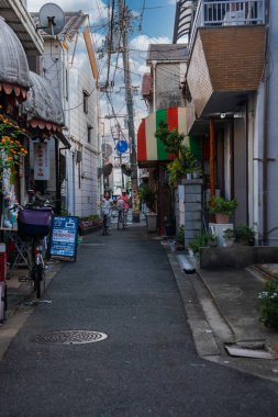 Dar bir Osaka sokağı küçük dükkanlar, saksı bitkileri ve Japon tabelalarını gösteriyor. İki yerli halk, güpegündüz elektrik kablolarının altında kameraya doğru bisiklet sürüyor..
