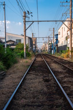 Zemin kat manzarası, elektrik direkleri, kateneri kabloları ve evleri olan bir Osaka mahallesi boyunca dar gösterge izleri gösteriyor. Öğleden sonra ışığı uzun gölgeler bırakır.