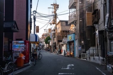 Sessiz bir Osaka caddesinde alçak katlı binalar, otomatlar, park edilmiş bisikletler ve çapraz elektrik hatları görülüyor. Yumuşak ışık, kepenkli dükkanlara ve mavi tenteye gölge düşürüyor..