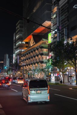 Osaka, Japonya 'da ışıklı bir cadde modern bir kulenin altında katmanlı bir cephenin önünden geçen arabaları, parlak yeşil tabelaları ve dikey çerçeveli bulvarı çevreleyen ağaçları gösteriyor..