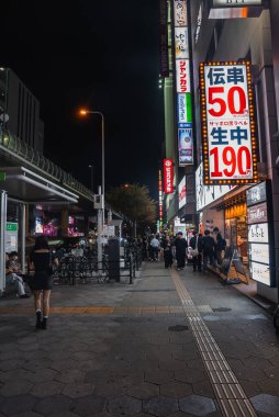 Dotonbori bölgesinde gece vakti Osaka caddesinde neon tabelalar, lokantalar, barlar ve park edilmiş bisikletler görülüyor. Yayalar dükkan önlerinden ve 50 ve 190 yen fiyatlı dikey tabelalardan geçiyor.