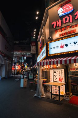 Gece Osaka Dotonbori 'deki dar sokak yemekhanelerinde Japon ve Koreli ışıklı tabelalar, ramen tabureleri ve menü tahtaları, sıcak renkler ve derin gölgeler.