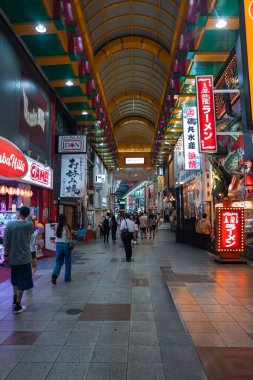 Osaka, Japonya 'da kapalı bir alışveriş merkezinde neon, fener işaretleri, ramen ve oyun reklamları gösteriliyor. Müşteriler ve lokantalar akşam ışığında sarı kemerli bir çatı altında yürürler..