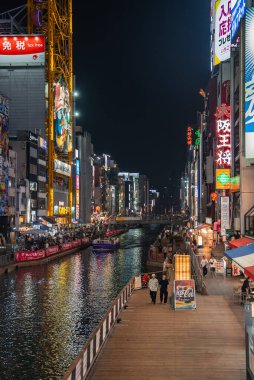 Dotonbori, Osaka 'daki gece sahnesinde neon panolar, reklam panoları ve yansımaları var. İnsanlar, Don Kişot cephesinin önünden kayarak geçerken tahta kaldırımlarda ve köprülerde yürüyor..