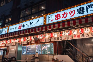 Osaka Shinsekai 'deki gece sokak sahnesinde beyaz ve kırmızı fenerler ve tabelalarla aydınlatılmış bir kushikatsu restoranı görülüyor. Menü tahtaları, çizgili tenteler ve sıcak tungsten ışıltısı var..