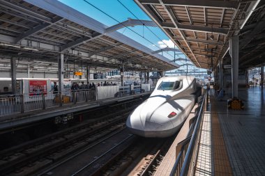 Beyaz bir Shinkansen, Japonya 'da kapalı bir platforma giriyor ve yolcular bariyerlerin arkasında bekliyor. Gün ortası ışığı kıtır gölgeler oluşturur, geniş açı görünümü modern geometriyi vurgular.