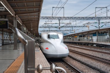 Kırmızı ışıklı beyaz bir Shinkansen Osaka istasyonuna giriyor. Bulutlu gün ışığı, izleri, işaretleri, gölgeleri ve trenin aerodinamik burnunu gösteren serin tonlar verir..