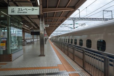 Beyaz bir Shinkansen yüksek bir Osaka platformunda durur, oval pencereler hizalanır, güvenlik korkulukları ve katenareri hatları görünür olur, bulutlu ışık altında Japonca ve İngilizce işaretler mevcuttur..