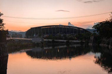 Osaka 'da modern bir oval stadyum alacakaranlıkta duruyor. Kaburgaları durgun sularda, alçak dağlarda, sakin bir deniz kıyısındaki patikadan seyrediliyor..