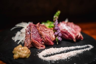 Koyu taş levhalarda kurutulmuş az pişmiş sığır eti, soğan, kaba tuz, yaban arısı, şiso çiçeği ve Osaka 'da mikro yeşillikler, sıcak ışık, sığ odak.