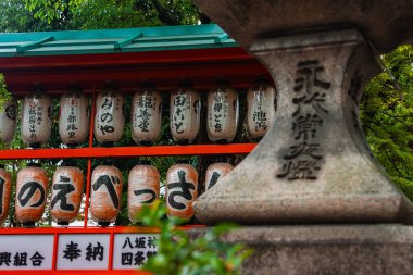 Siyah kanjili geleneksel kağıt fenerler Kyoto 'da mercan çatı altında asılı ve kırmızı kirişler altında. Bitkiler ve bulutlu ışık tarafından çerçevelenmiş yıpranmış taş bir fener önünde duruyor..