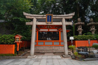 Kyoto 'da bir Shinto tapınağı mavi plakalı granit bir toridinin arkasında duruyor. Taş toro fenerleri temiz bir yolu kuşatır. Yumuşak gün ışığı ve ön simetri dikkatli bir zanaat gösteriyor.