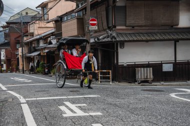 Bir triportör, Japonya 'nın Kyoto kentindeki Machiya konakları boyunca iki yolcuyla birlikte bir jinrikisha çekiyor. Bulutlu gün ışığı kiremitli çatılar, kafes pencereler ve taş bir yol gösteriyor..