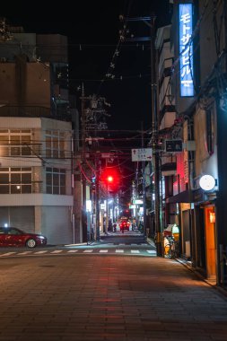 Gece Kyoto 'nun yan sokağında kırmızı ışık, dikey kanji neonu, alçak cepheler, elektrik hatları, boş bir yaya geçidi, park halindeki kırmızı bir araba ve fener ışığında yanan bir kapı bulunur..