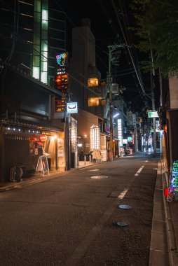 Geceleri dar bir Kyoto caddesinde küçük restoranlar ve barlar, sıcak fenerler, neon lambaları, yeşil bir eczane işareti, elektrik hatları ve seyrek yaya trafiği olan yüksek binalar görülüyor..