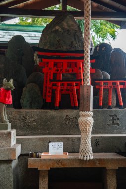 Kyoto 'daki küçük Shinto türbesi sunağı istiflenmiş vermilion torii, taş muhafızlar, kırmızı önlüklü tilki heykeli, çan ipi ve sunucu kutusu yumuşak gün ışığı altında ve sıkı çerçeveli.