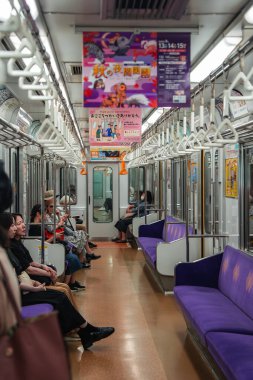 Bir Kyoto, Japon metro vagonu mor banklarda, el kayışlarında ve çelik direklerde oturan binicileri gösteriyor, posterler ve güzergah uyarıları yumuşak floresan ışıkların altında asılı..