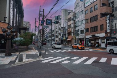 Japonya 'da bir Nara caddesi alacakaranlıkta, yükselen binaların ortasında Japon tabelaları, elektrik hatları yukarıda, arabalar ve turuncu bir taksi geniş bir zebra geçidini geçiyor, saksı bitkilerinin metal sanat eserleri..
