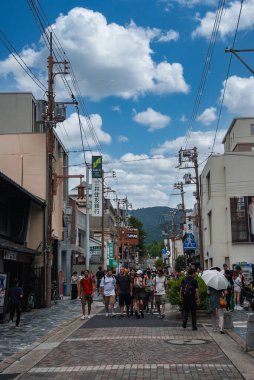 Japonya, Nara 'da yoğun bir şeritte şemsiyeli ve kameralı turistler, tahta direkler, dükkan önleri, Japon tabelaları, mavi bir geçiş işareti ve yazın uzak tepeler görülüyor.